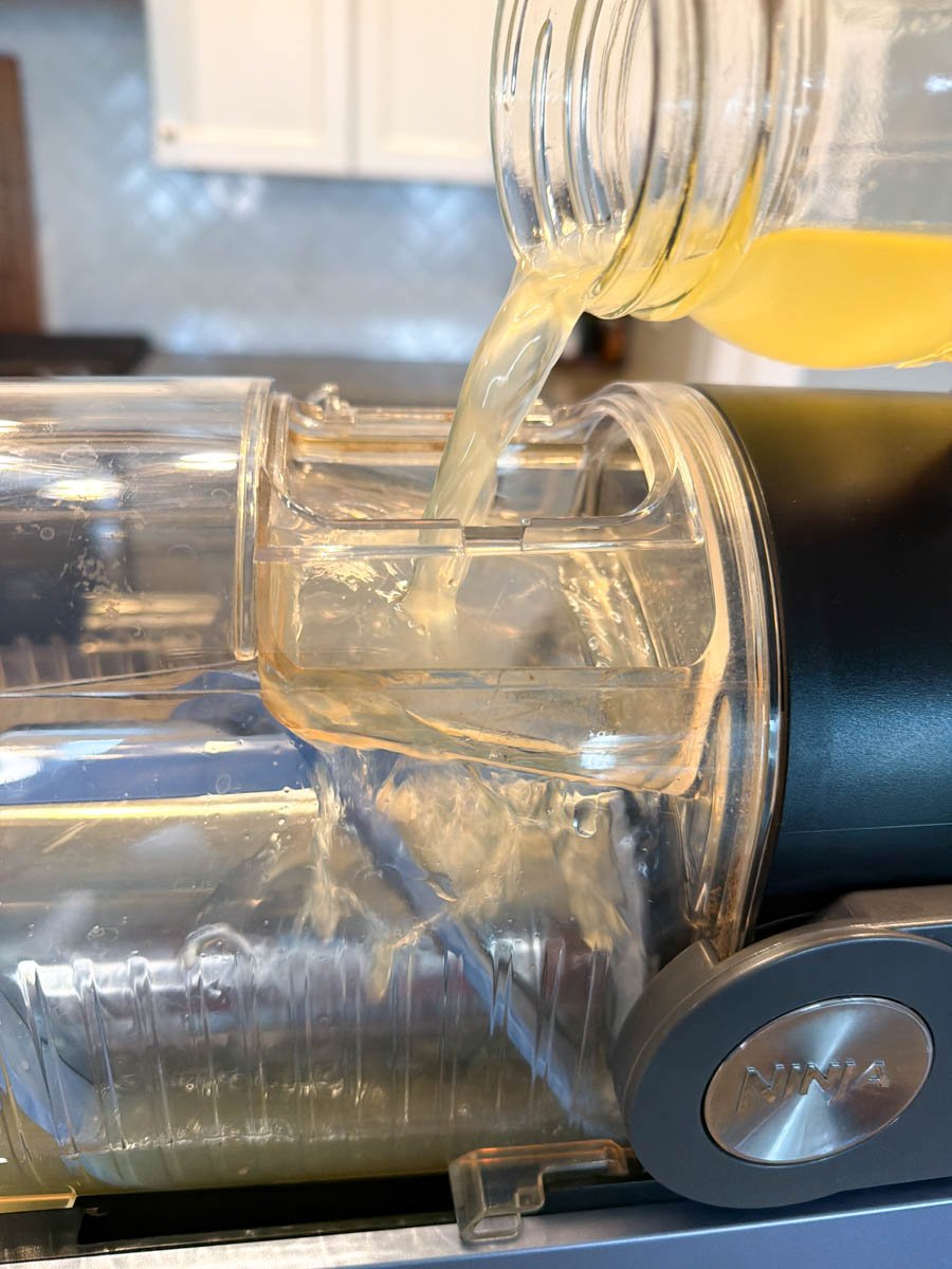 A jar of orange cocktail mixture being poured into the top of the Ninja Slushi machine, creating a flowing stream into the frozen base inside.