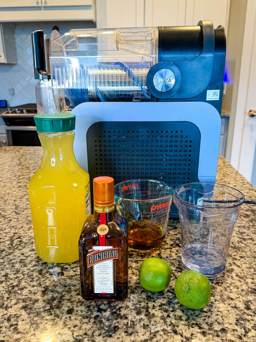 A countertop setup with the Ninja Slushi machine and ingredients arranged in front: a bottle of juice, a bottle of Cointreau, a measuring cup of dark rum, an empty cup, and two whole limes.