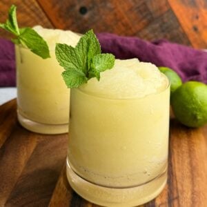 Two glasses of pale yellow frozen mojito slush garnished with fresh mint sprigs, sitting on a wooden board with whole limes and a purple cloth in the background.