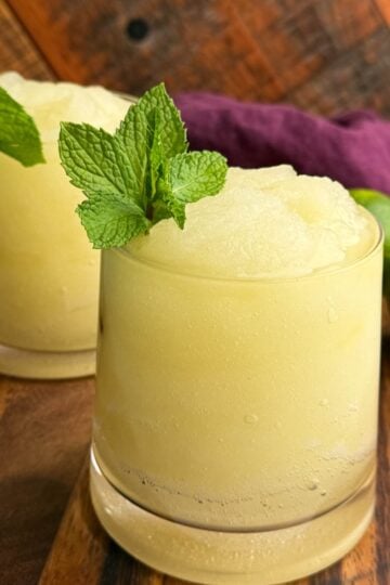 Two glasses of pale yellow frozen mojito slush garnished with fresh mint sprigs, sitting on a wooden board with whole limes and a purple cloth in the background.