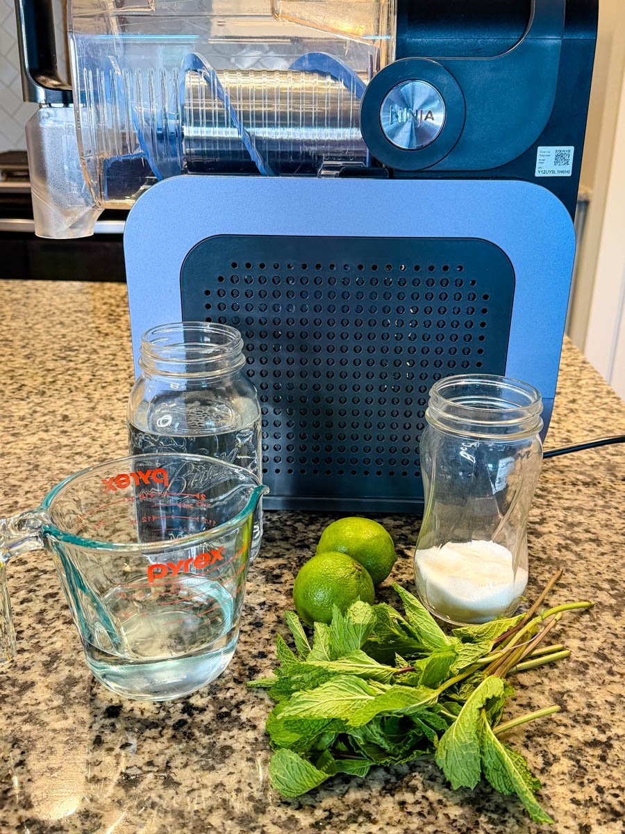 A countertop setup with a Ninja slushi machine in the background and mojito ingredients arranged in front: two empty glass jars, a measuring cup filled with water, two whole limes, a bunch of fresh mint, and a small jar of sugar.