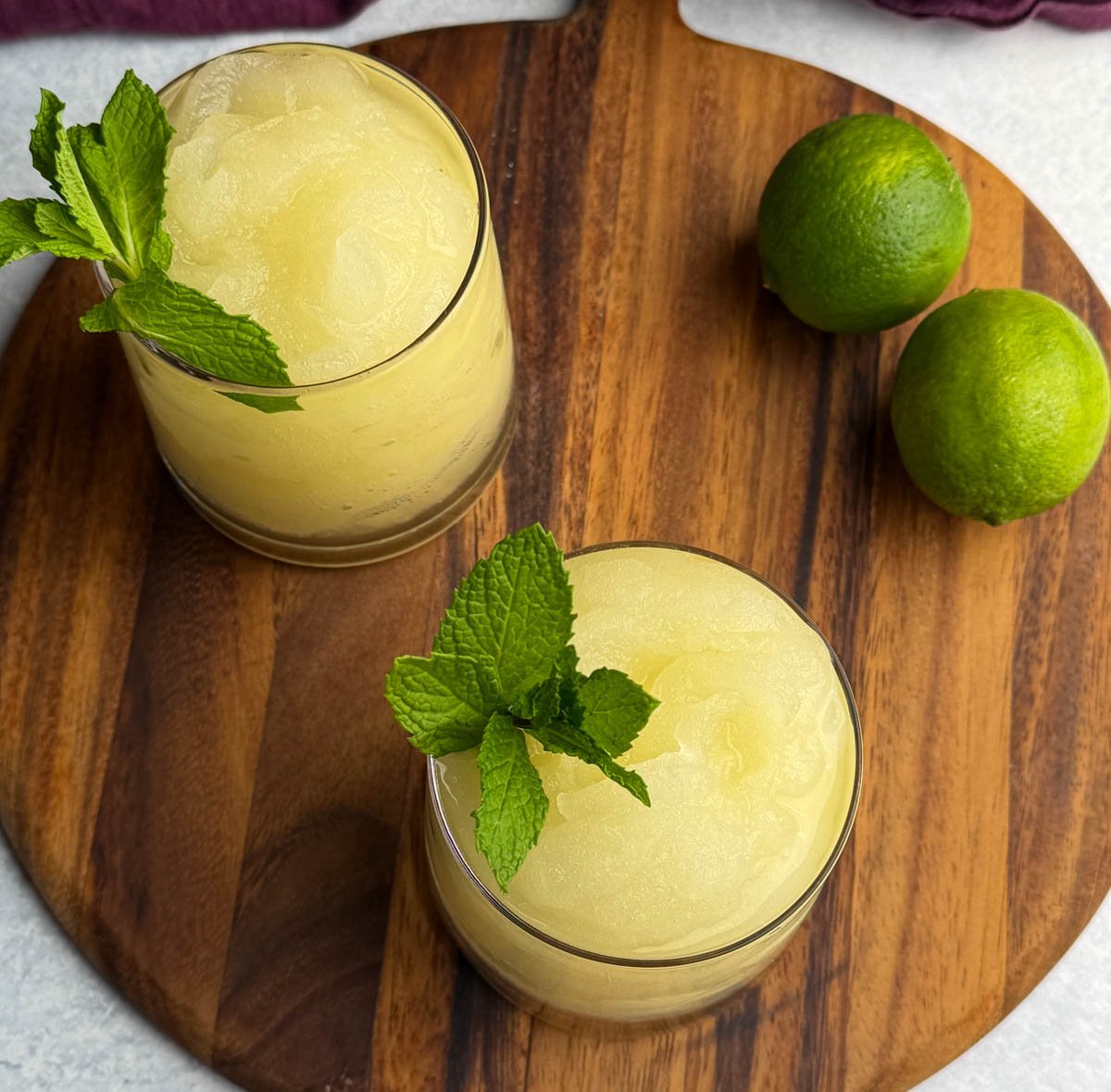 An overhead view of two glasses of frozen mojito slush garnished with fresh mint. The glasses are placed on a round wooden board with whole limes off to the side.
