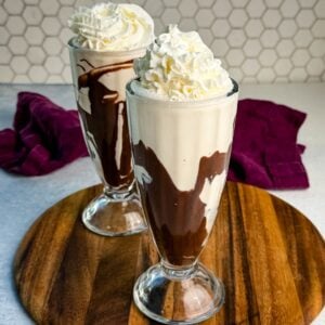 Two tall glasses filled with creamy Ninja Slushi Mudslides swirled with chocolate syrup and topped with whipped cream, placed on a wooden board with a purple cloth in the background.