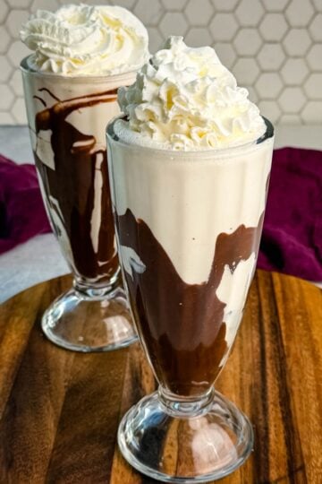 Two tall glasses filled with creamy Ninja Slushi Mudslides swirled with chocolate syrup and topped with whipped cream, placed on a wooden board with a purple cloth in the background.