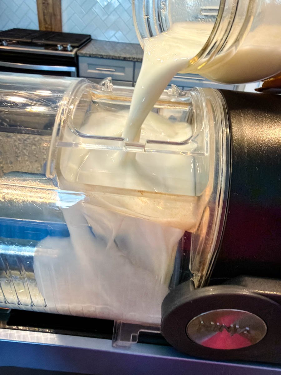 Creamy mudslide mixture being poured from a jar into the Ninja Creami container, showing the smooth liquid filling up the machine insert with a kitchen stove and backsplash in the background.