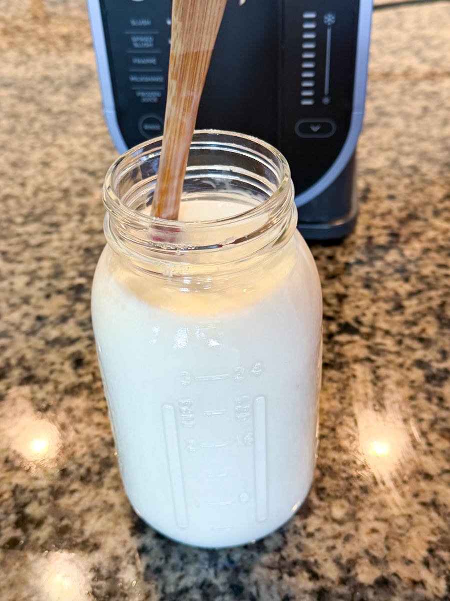 A glass mason jar filled with creamy mudslide mixture and a wooden spoon inside, sitting on a granite countertop in front of a Ninja Creami machine.