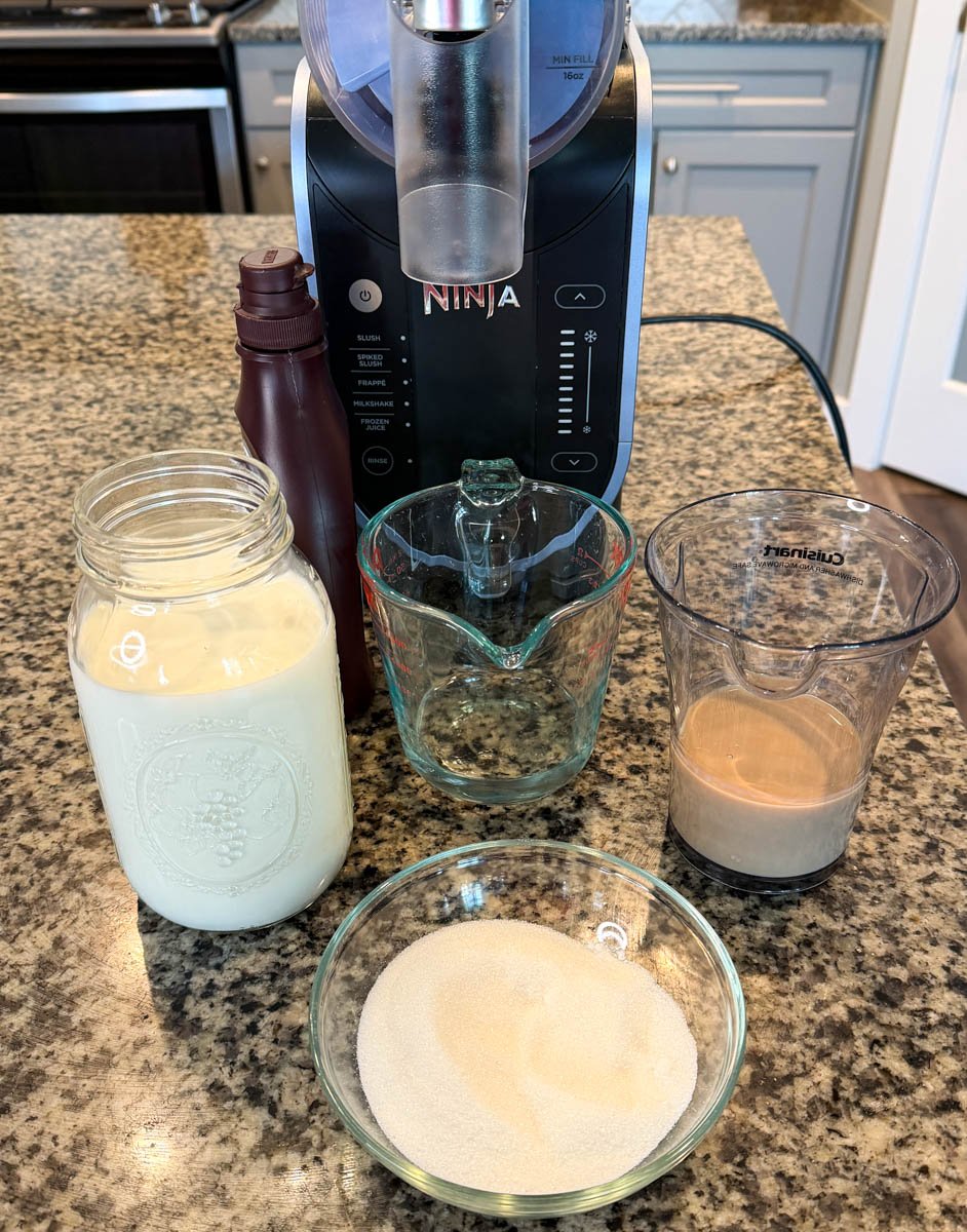 Ingredients for a Ninja Slushi Mudslide arranged on a countertop, including a jar of milk mixture, a small bowl of sugar, a measuring cup with Irish cream, and chocolate syrup in front of the Ninja Creami machine.