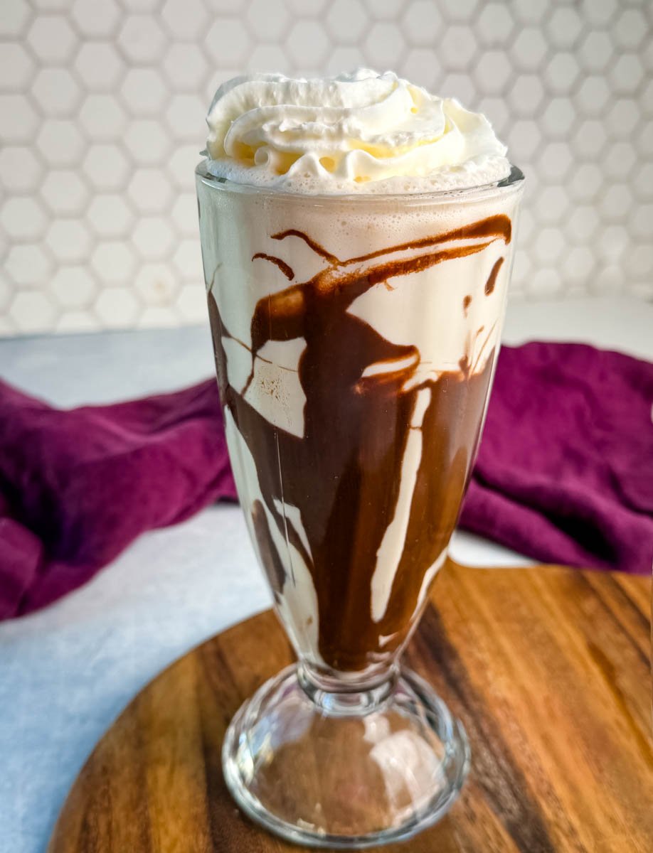 Close-up of one tall glass filled with a Ninja Slushi Mudslide. The glass is drizzled with chocolate syrup and topped with whipped cream, sitting on a wooden board with a hexagon-tile backsplash in the background.