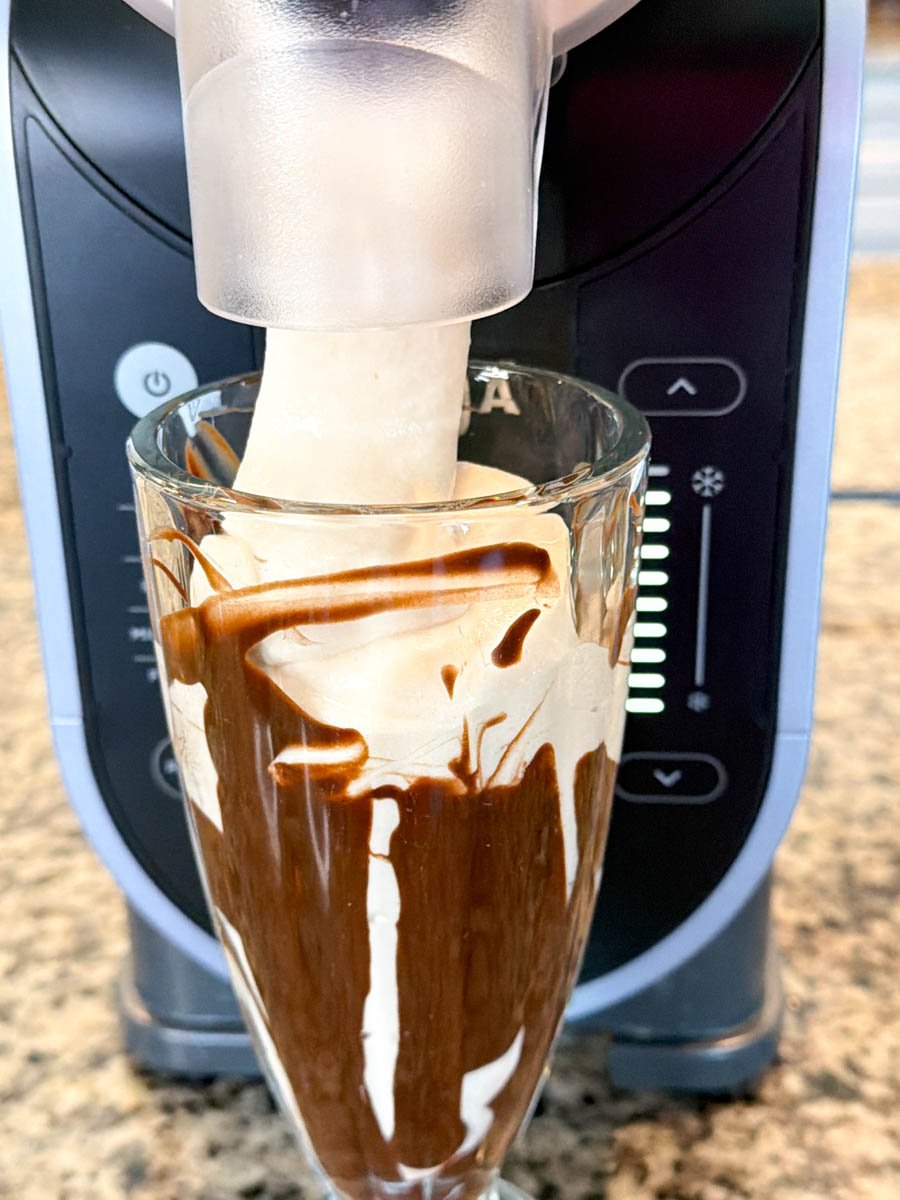 Close-up of a Ninja Creami machine dispensing a thick, creamy mudslide mixture into a tall glass lined with chocolate syrup, showing the texture as it pours out smoothly.
