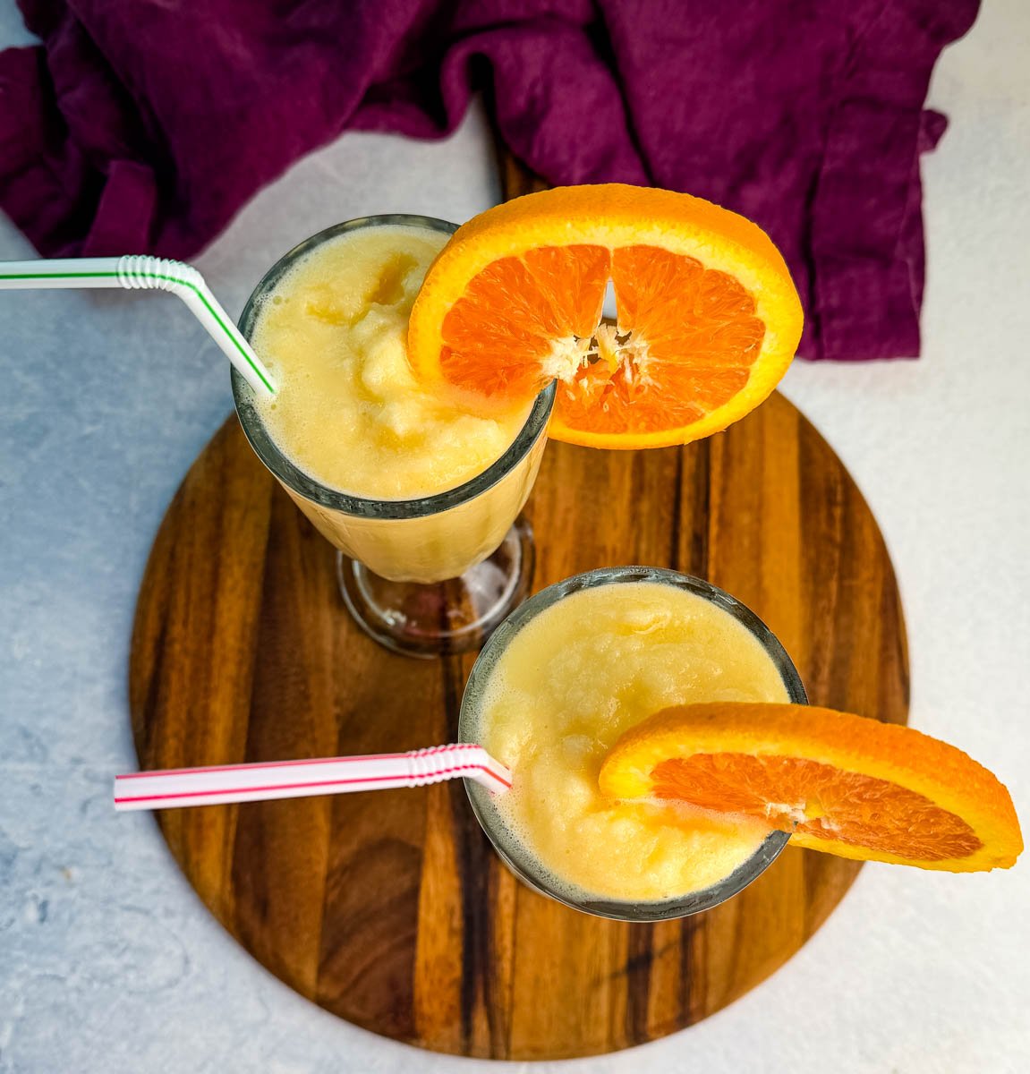 Two glasses of frozen orange Julius slush topped with orange slices and straws, set on a wooden serving board with a purple cloth in the background.