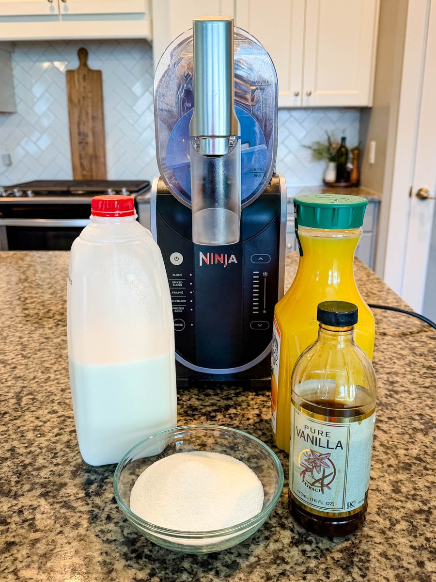 Ingredients for a Ninja Slushi Orange Julius displayed on a granite countertop, including a bottle of milk, a bowl of sugar, a bottle of vanilla extract, and a bottle of orange juice, with the Ninja Slushi machine positioned behind them in a bright kitchen.