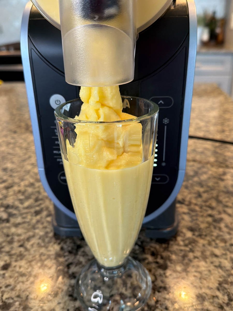 A thick, creamy orange Julius slushie dispensing from the Ninja Slushi machine into a tall glass on a granite countertop.