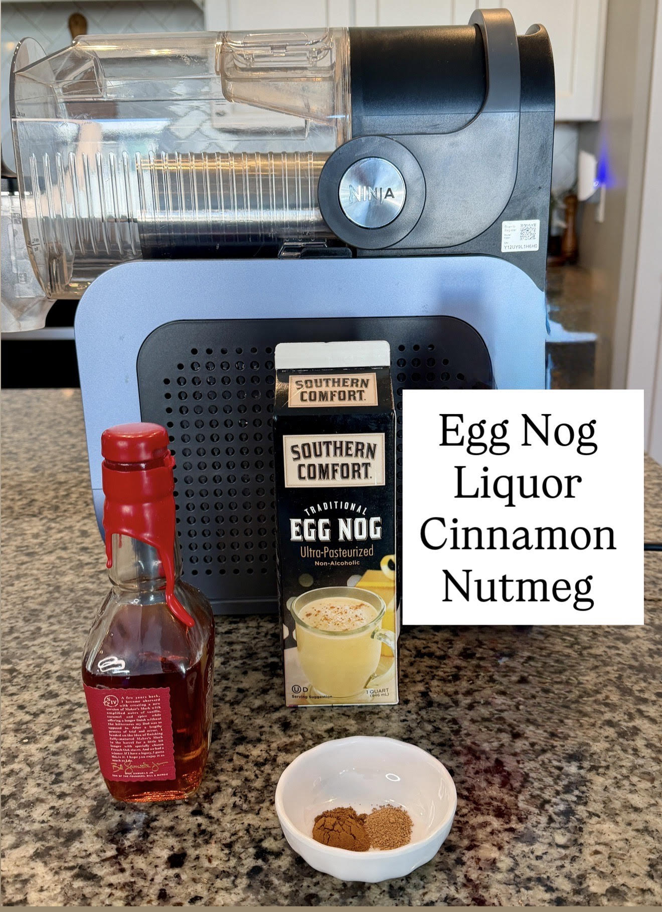 Ingredients for the spiked eggnog recipe arranged in front of a Ninja Slushi machine, including Southern Comfort eggnog, a bottle of bourbon, whipped topping, and a small bowl of spices.
