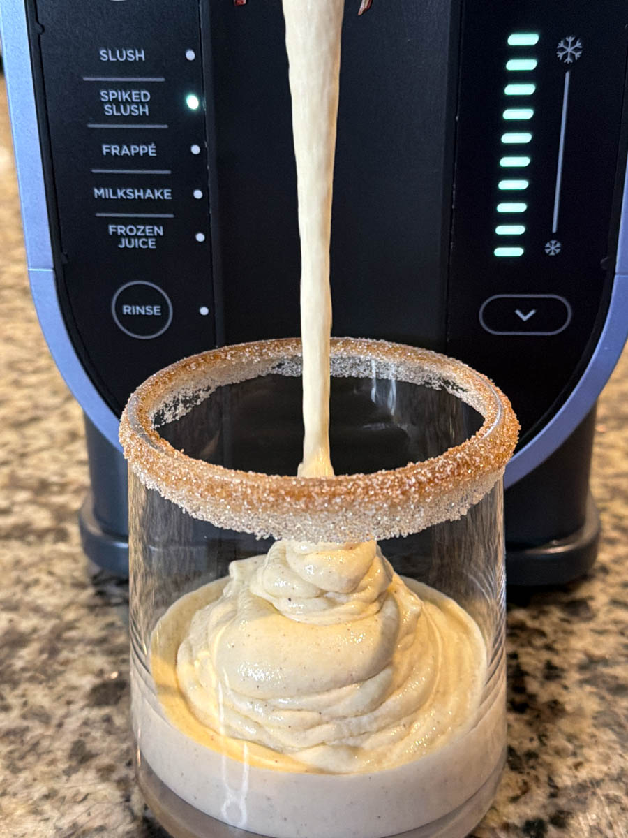 Thick, creamy spiked eggnog being dispensed from the Ninja Slushi machine into a glass with a cinnamon sugar rim.