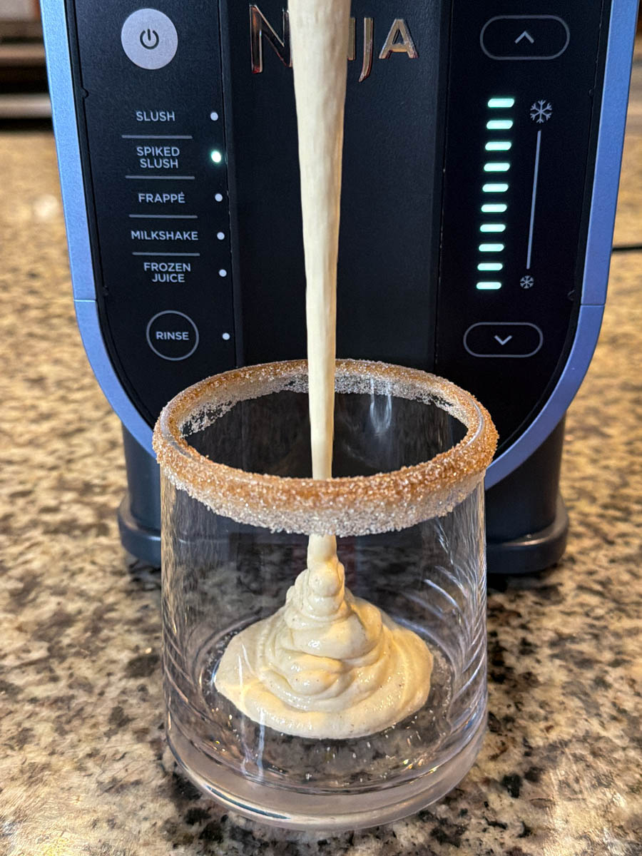 Thick, creamy spiked eggnog being dispensed from the Ninja Slushi machine into a glass with a cinnamon sugar rim.
