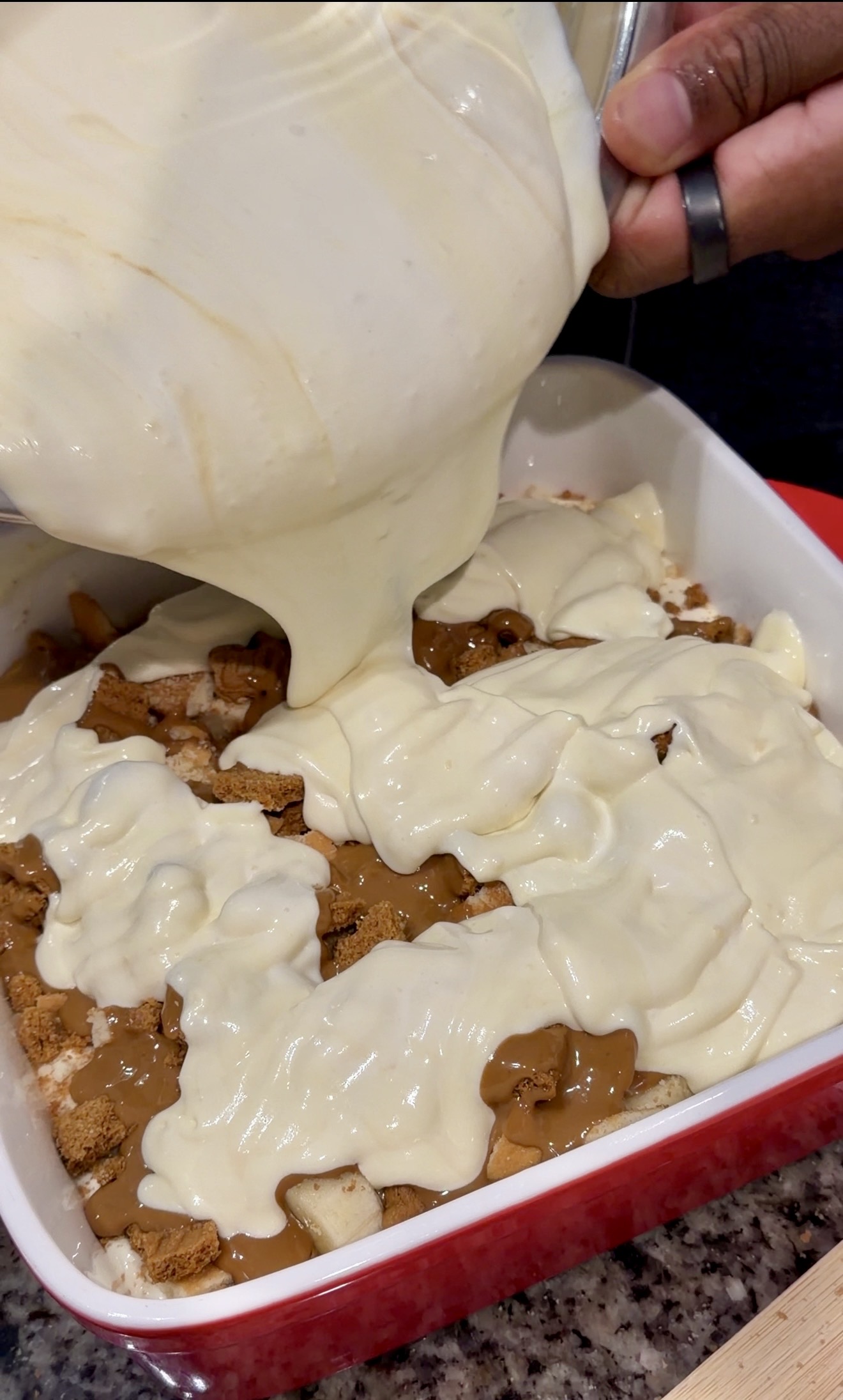Creamy banana pudding mixture being poured over layers of bananas, crushed Biscoff cookies, and cookie butter in a baking dis