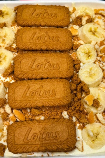 Overhead view of Biscoff banana pudding layered with sliced bananas, crushed Biscoff cookies, and whole Lotus Biscoff biscuits arranged on top in a white baking dish.