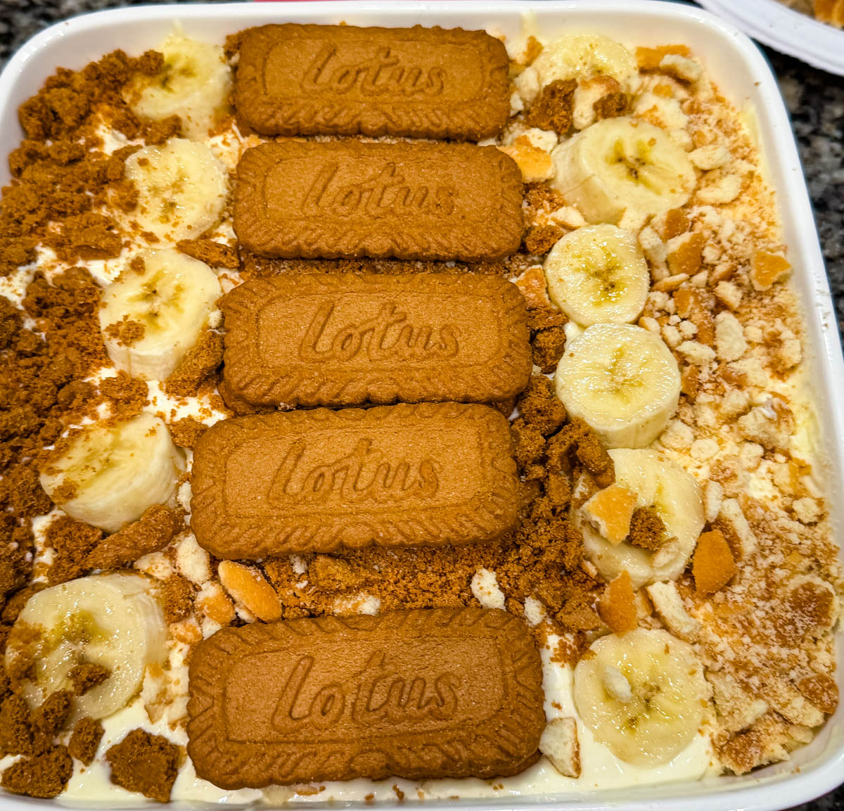 Overhead view of Biscoff banana pudding layered with sliced bananas, crushed Biscoff cookies, and whole Lotus Biscoff biscuits arranged on top in a white baking dish.