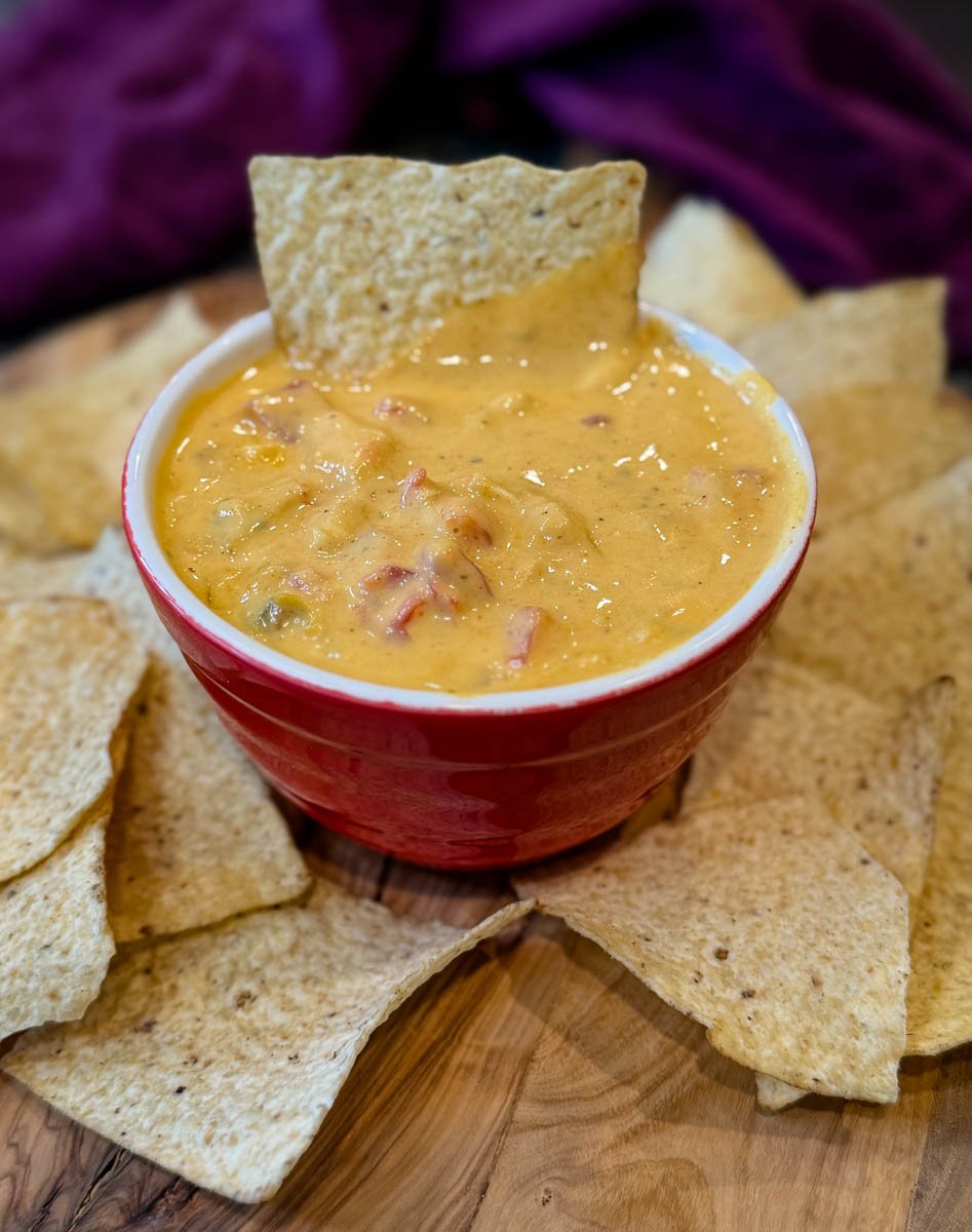 Bowl of creamy Chicken Rotel Dip without Velveeta topped with a tortilla chip and surrounded by additional tortilla chips on a wooden board.