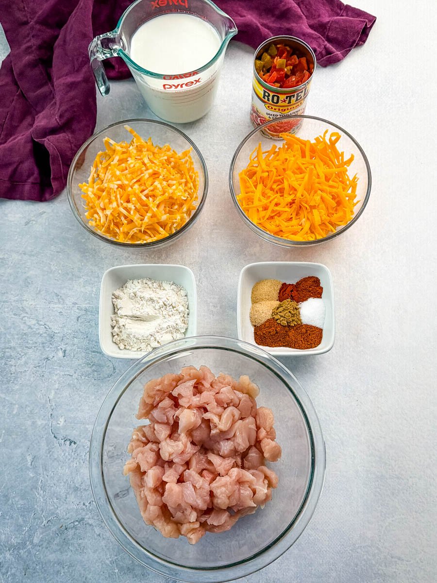 Ingredients for Chicken Rotel Dip without Velveeta displayed on a countertop, including diced chicken, shredded cheddar and Colby Jack cheese, flour, spices, Rotel tomatoes and green chiles, and a measuring cup of half and half.