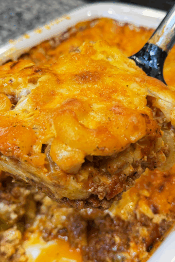 Close-up of a slice of macaroni and cheese lasagna lifted out of a baking dish, showing creamy elbows, meat sauce, and melted browned cheese.