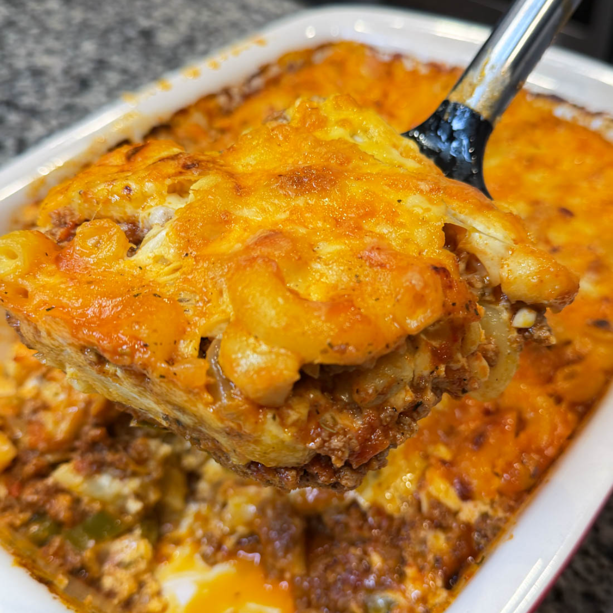 Close-up of a slice of macaroni and cheese lasagna lifted out of a baking dish, showing creamy elbows, meat sauce, and melted browned cheese.