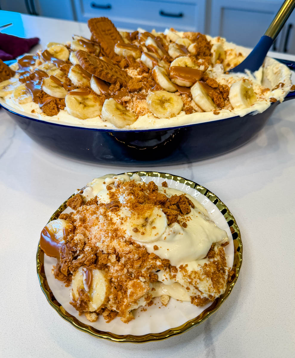 Served scoop of Biscoff banana pudding topped with bananas, crushed cookies, and cookie butter, with full dish in the background.