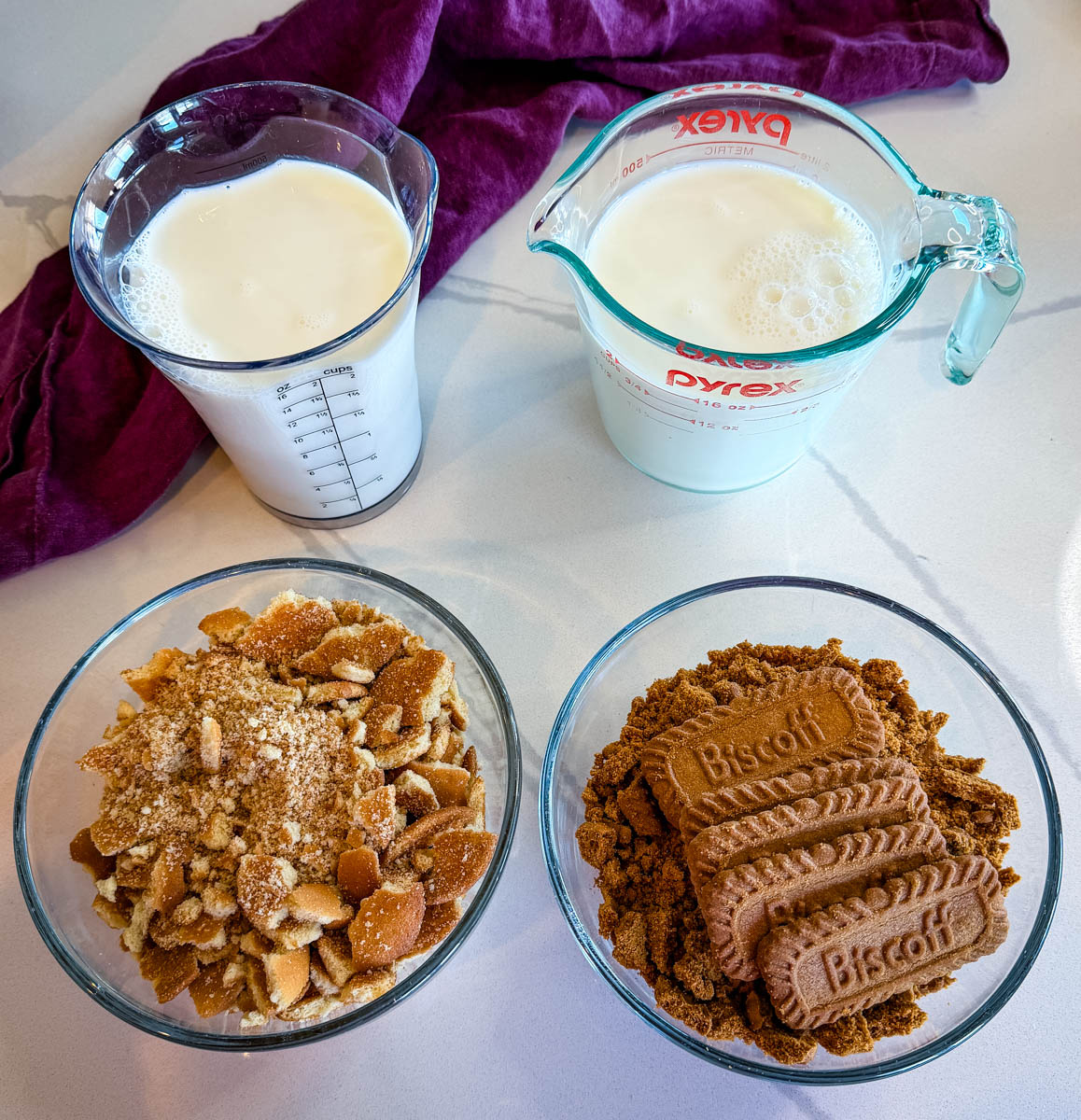 Measured milk in glass containers alongside bowls of crushed vanilla wafers and Biscoff cookies on a countertop.