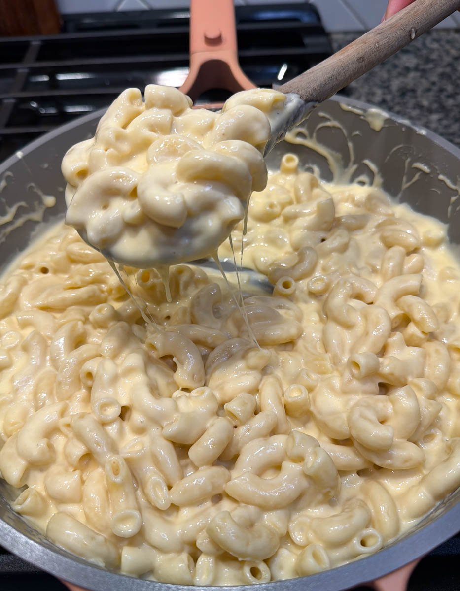 Creamy stovetop macaroni and cheese with elbows pasta coated in melted cheese sauce being lifted from a skillet with a wooden spoon.
