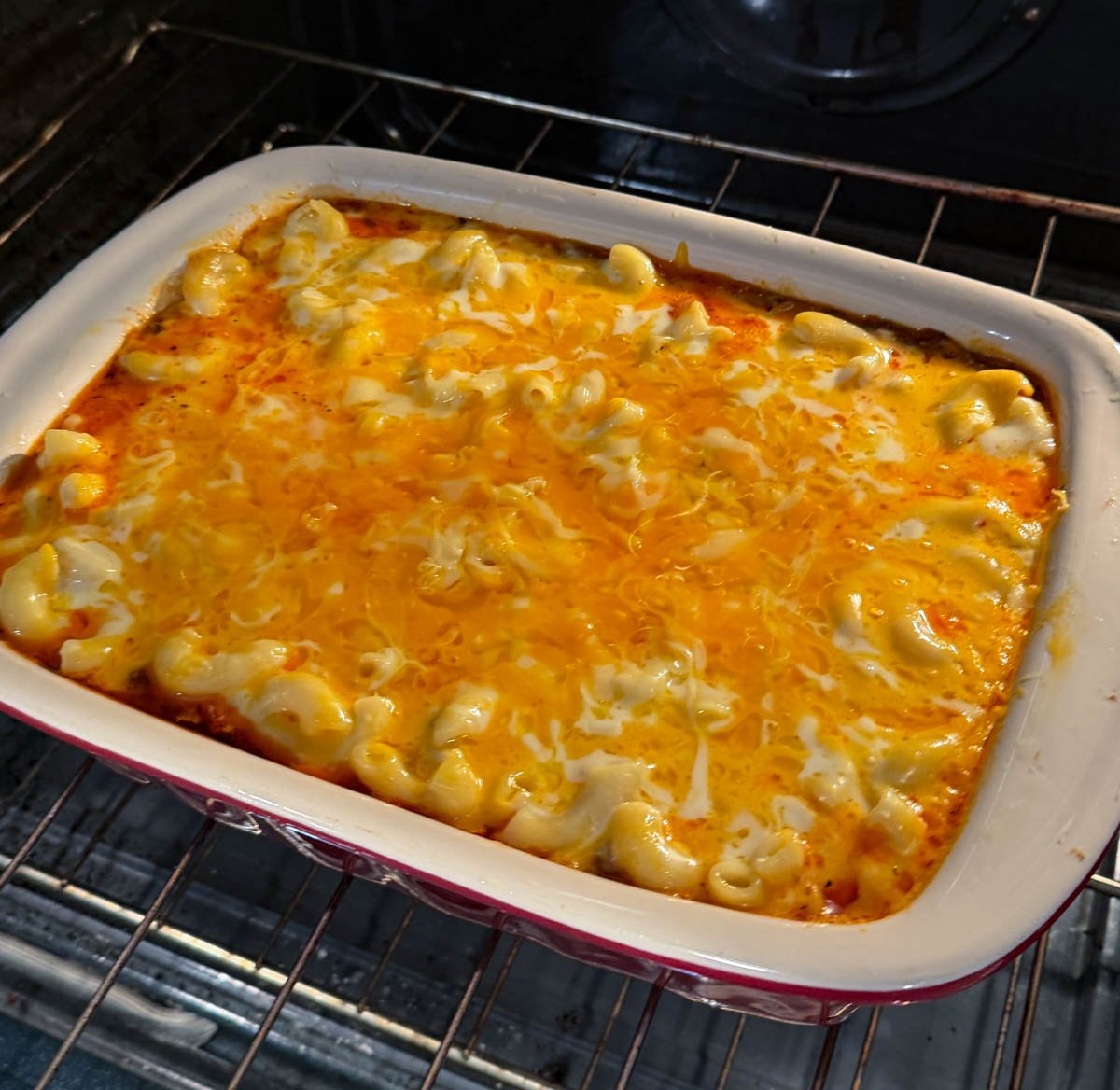 Macaroni and cheese lasagna in a red casserole dish baking in the oven with cheese melted and beginning to bubble but not yet browned.