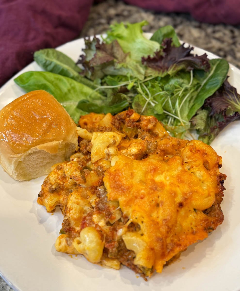 Serving of macaroni and cheese lasagna on a dinner plate with a green side salad and dinner roll.