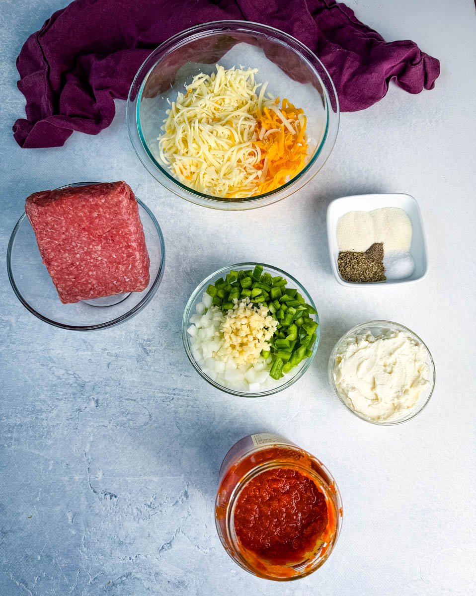 Overhead view of ground beef, shredded cheese, diced onions, green bell peppers, minced garlic, ricotta cheese, marinara sauce, and seasonings arranged in bowls on a light surface.