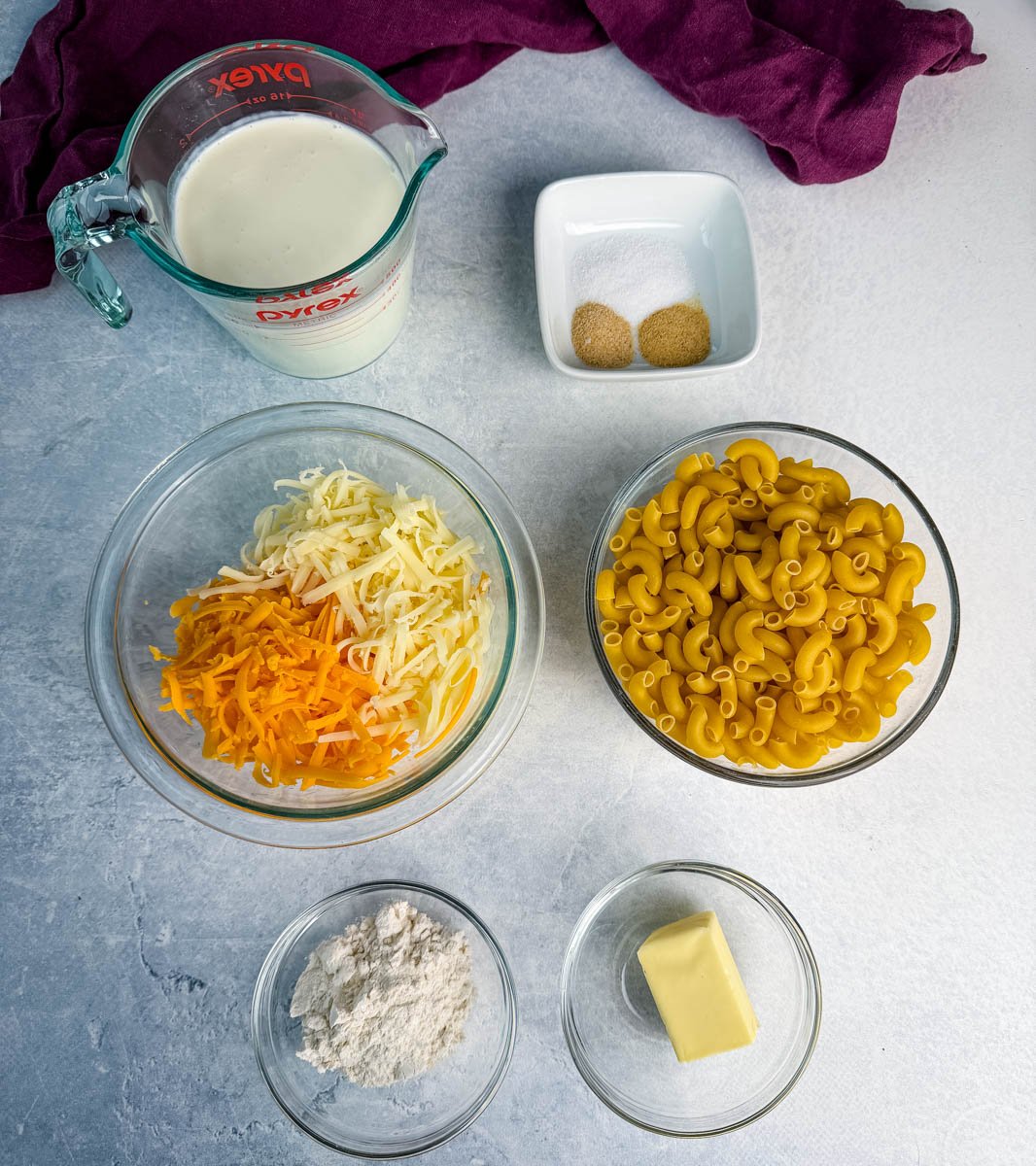Overhead view of elbows pasta, shredded cheeses, flour, butter, milk and cream mixture, and seasonings arranged in bowls on a light surface.