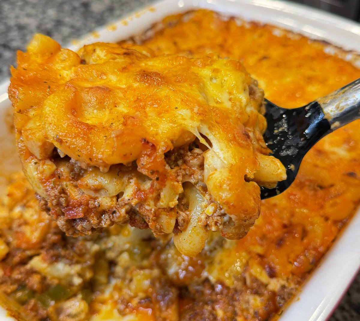 Close-up of a cheesy slice of macaroni and cheese lasagna lifted out of a casserole dish, showing browned cheese, elbows pasta, and meat sauce layers.