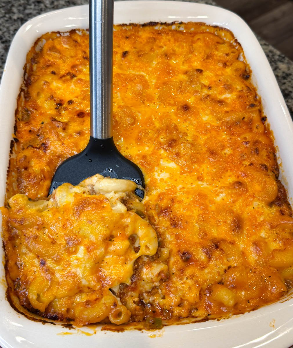 op-down view of baked macaroni and cheese lasagna in a casserole dish with a serving spoon lifting out a portion, showing golden brown cheesy top.