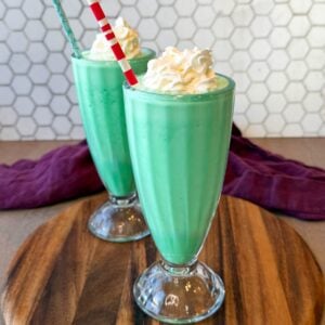 Mint green shamrock shakes topped with whipped cream and green sprinkles in soda fountain glasses.