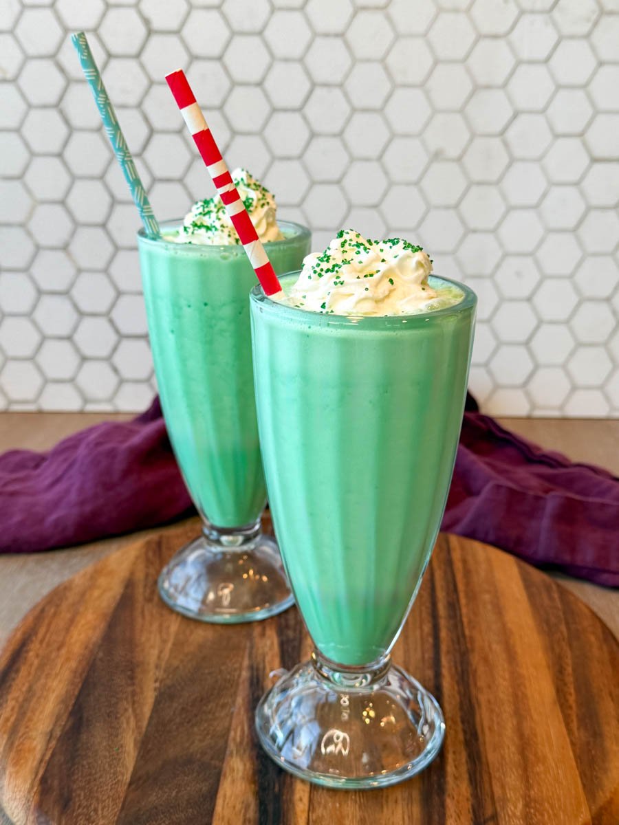 Two mint green shamrock shakes topped with whipped cream in tall glasses on a wooden board.