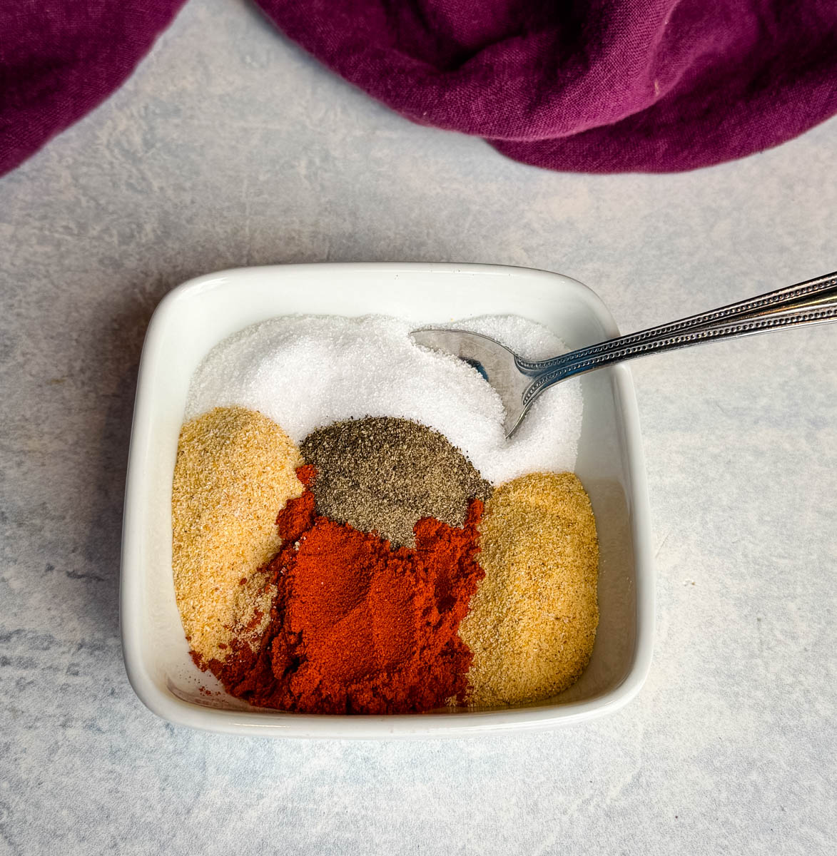 Overhead view of a small white bowl filled with separated spices including paprika, garlic powder, onion powder, salt, and black pepper with a spoon, ready to be mixed into homemade all purpose seasoning.