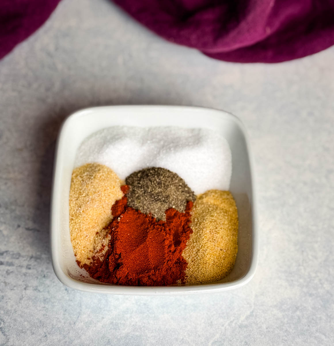 Overhead view of individual spices including paprika, garlic powder, onion powder, salt, and black pepper arranged in a small white bowl before mixing.