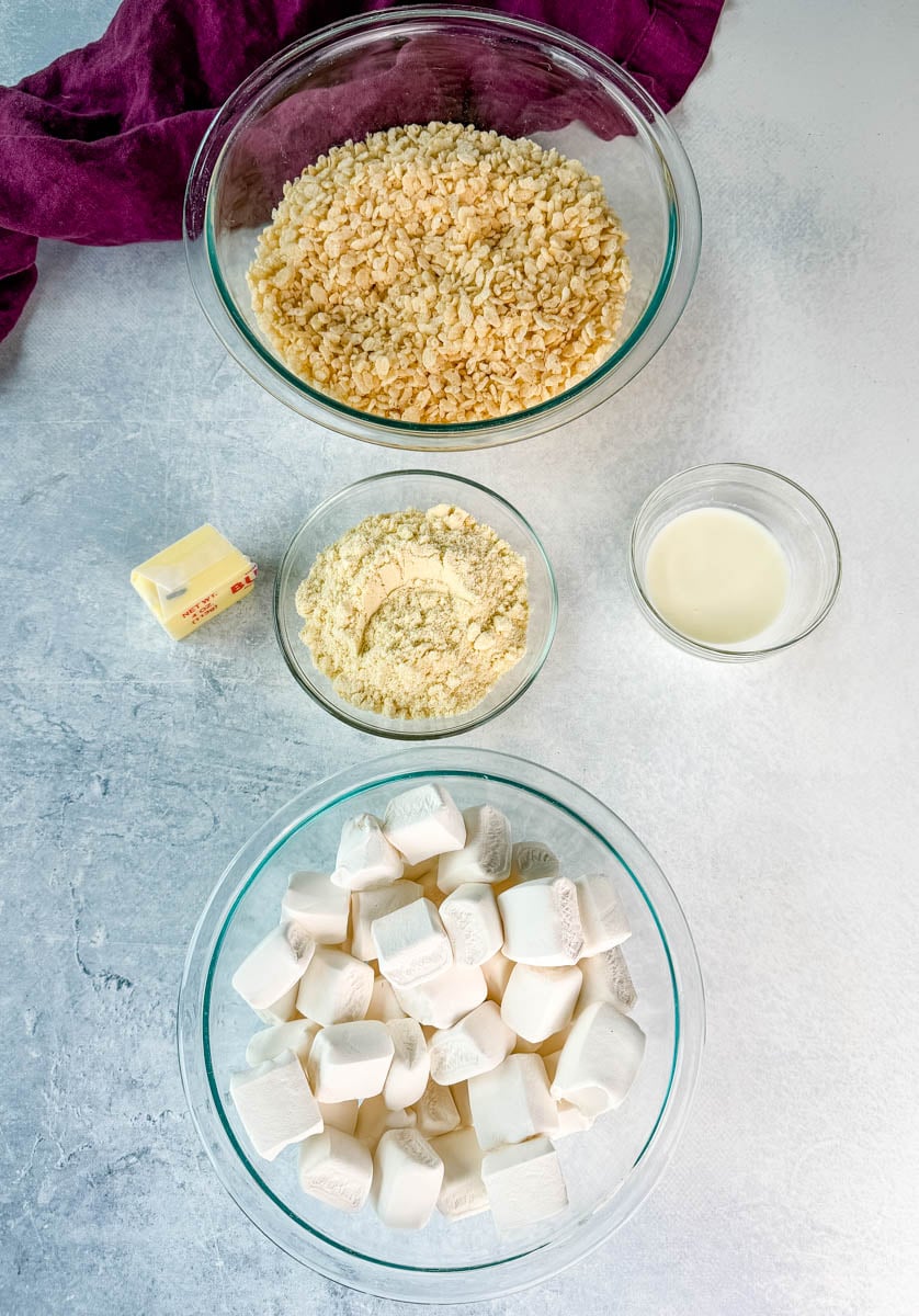 Ingredients for protein Rice Krispie treats including cereal, marshmallows, protein powder, butter, milk, and vanilla arranged in glass bowls.