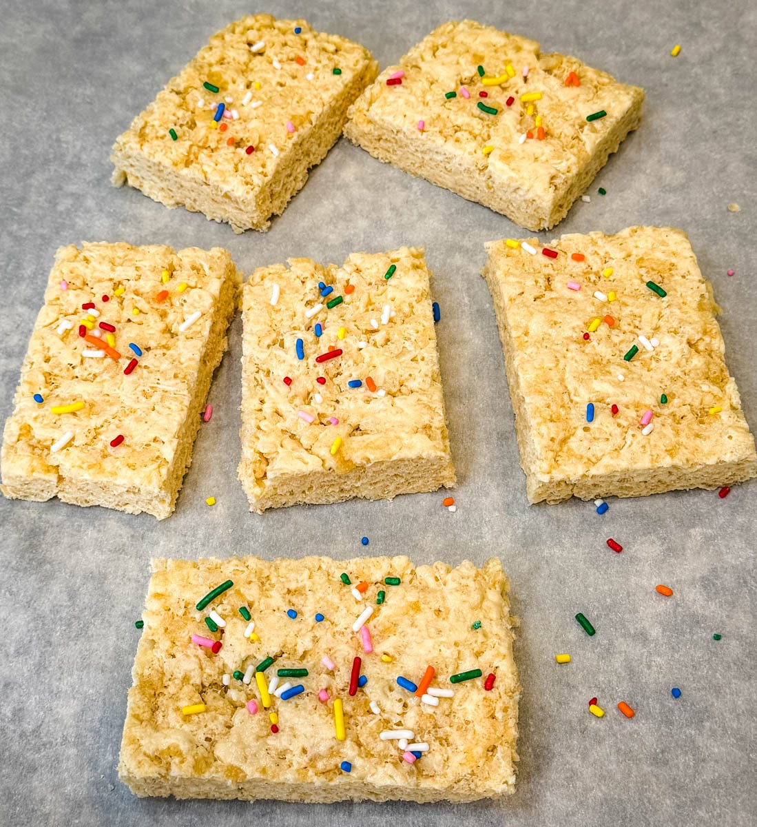 Protein Rice Krispie treats cut into squares and topped with rainbow sprinkles on parchment paper.