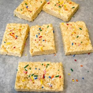Protein Rice Krispie treats cut into squares and topped with rainbow sprinkles on parchment paper.