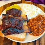 Oven baked beef back ribs served with baked beans, coleslaw, pickles, and sliced white bread on a plate.
