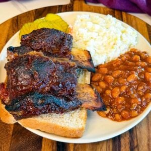 Oven baked beef back ribs served with baked beans, coleslaw, pickles, and sliced white bread on a plate.