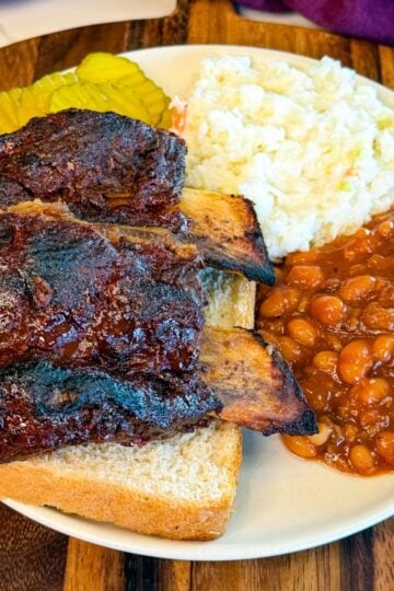 Oven baked beef back ribs served with baked beans, coleslaw, pickles, and sliced white bread on a plate.