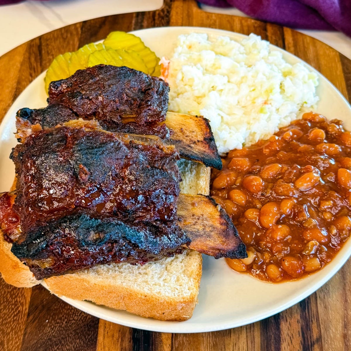 Oven baked beef back ribs served with baked beans, coleslaw, pickles, and sliced white bread on a plate.