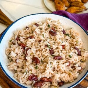 Bowl of Jamaican rice and peas with red kidney beans, coconut rice, and fresh herbs served on a wooden board with fried plantains in the background.