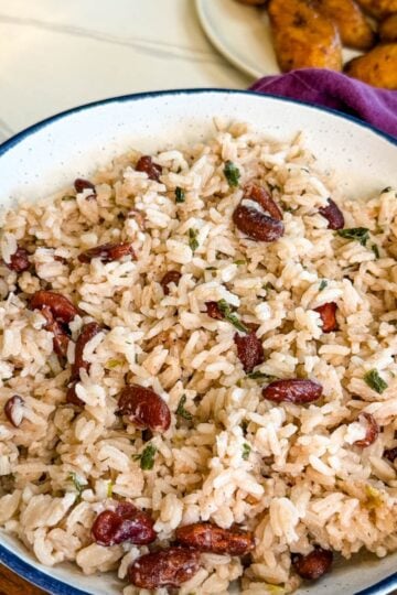 Bowl of Jamaican rice and peas with red kidney beans, coconut rice, and fresh herbs served on a wooden board with fried plantains in the background.