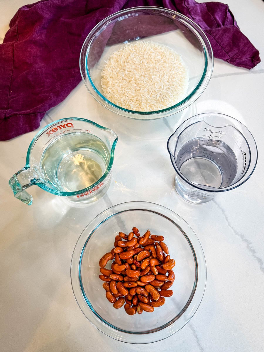 Uncooked rice, cooked red kidney beans, water, and coconut milk measured in bowls for Jamaican rice and peas.