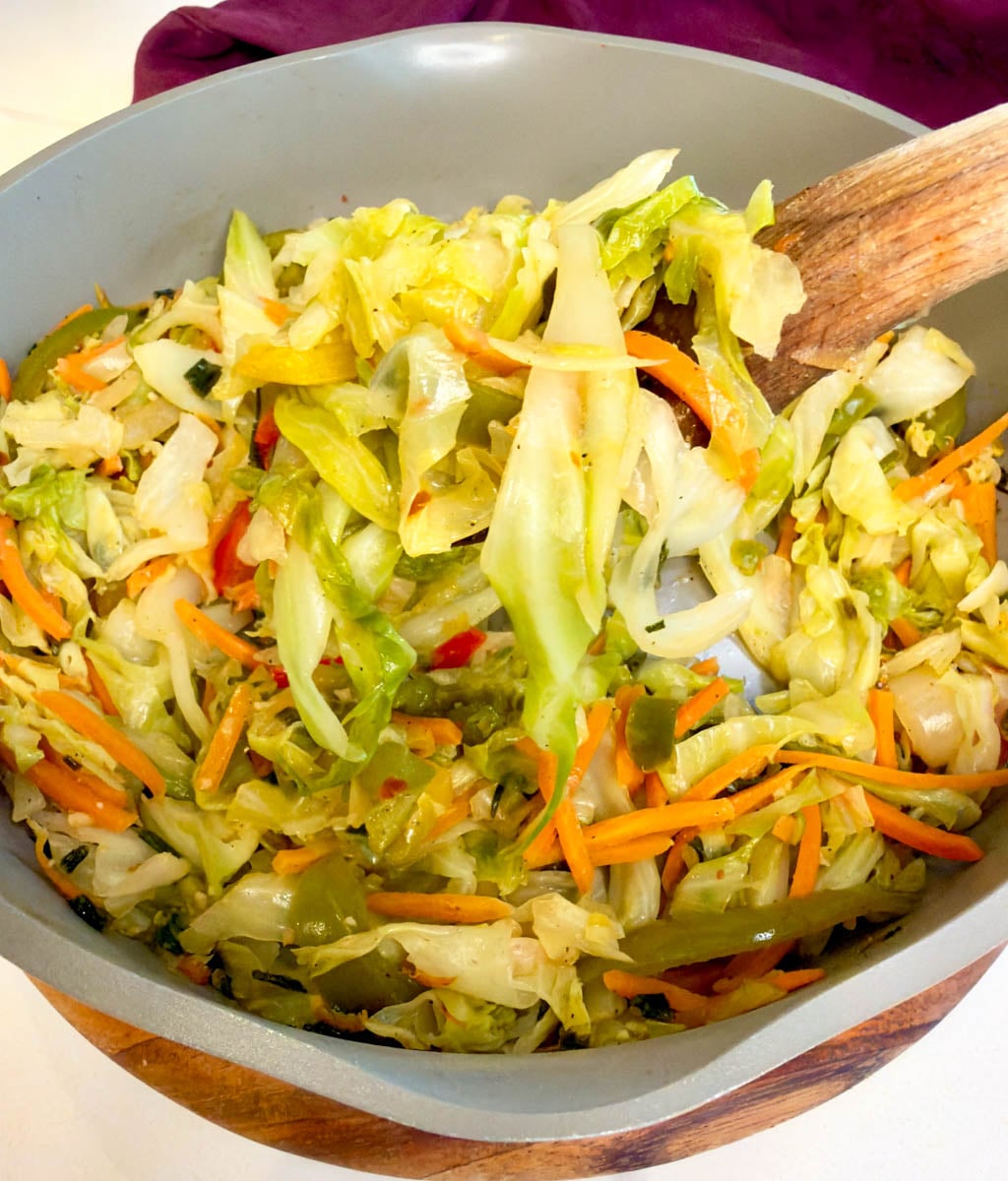 Jamaican steamed cabbage sautéing in a skillet with bell peppers, carrots, onions, and herbs being stirred with a wooden spoon.